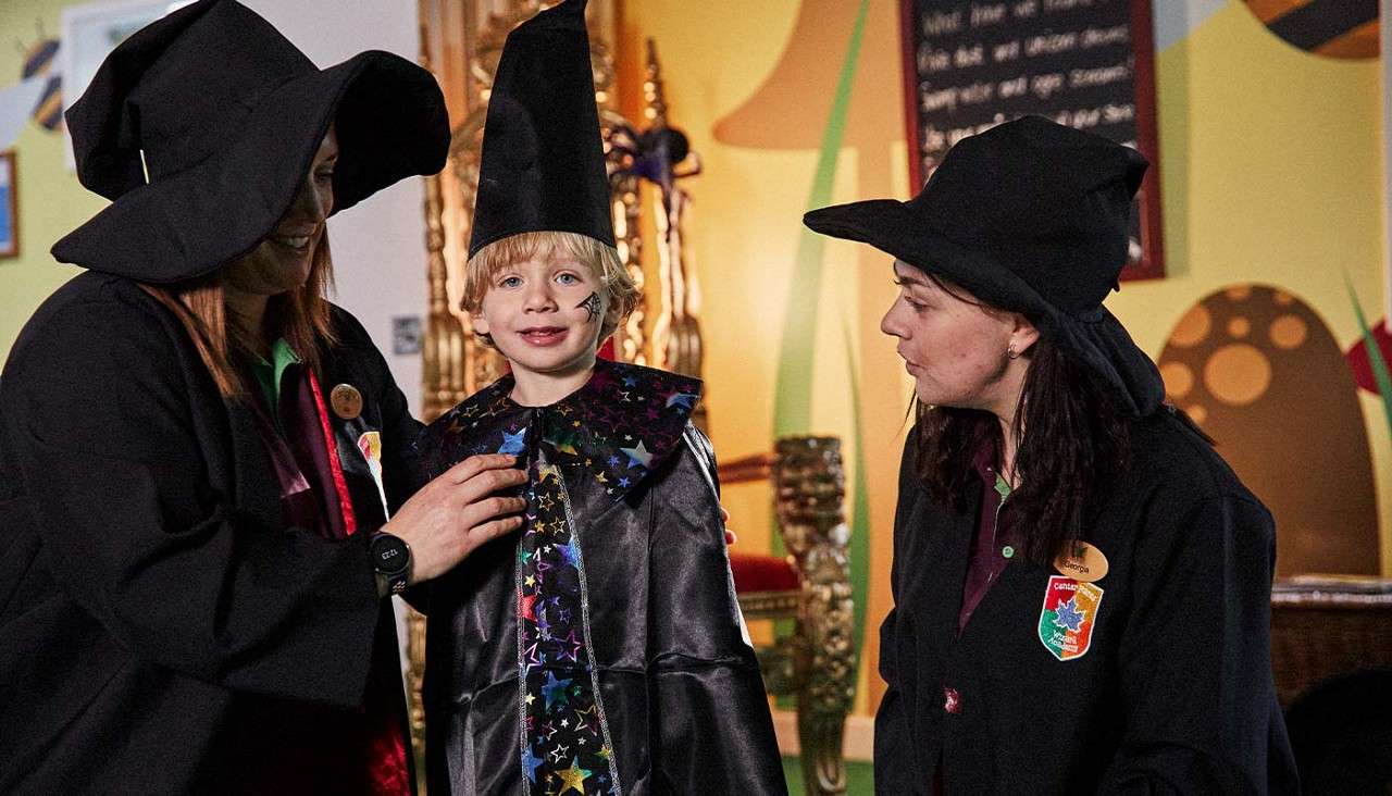 Child in wizard costume smiles as two adults in witch hats adjust the outfit, inside a colorful room with a gold throne and chalkboard. Text: Georgia.