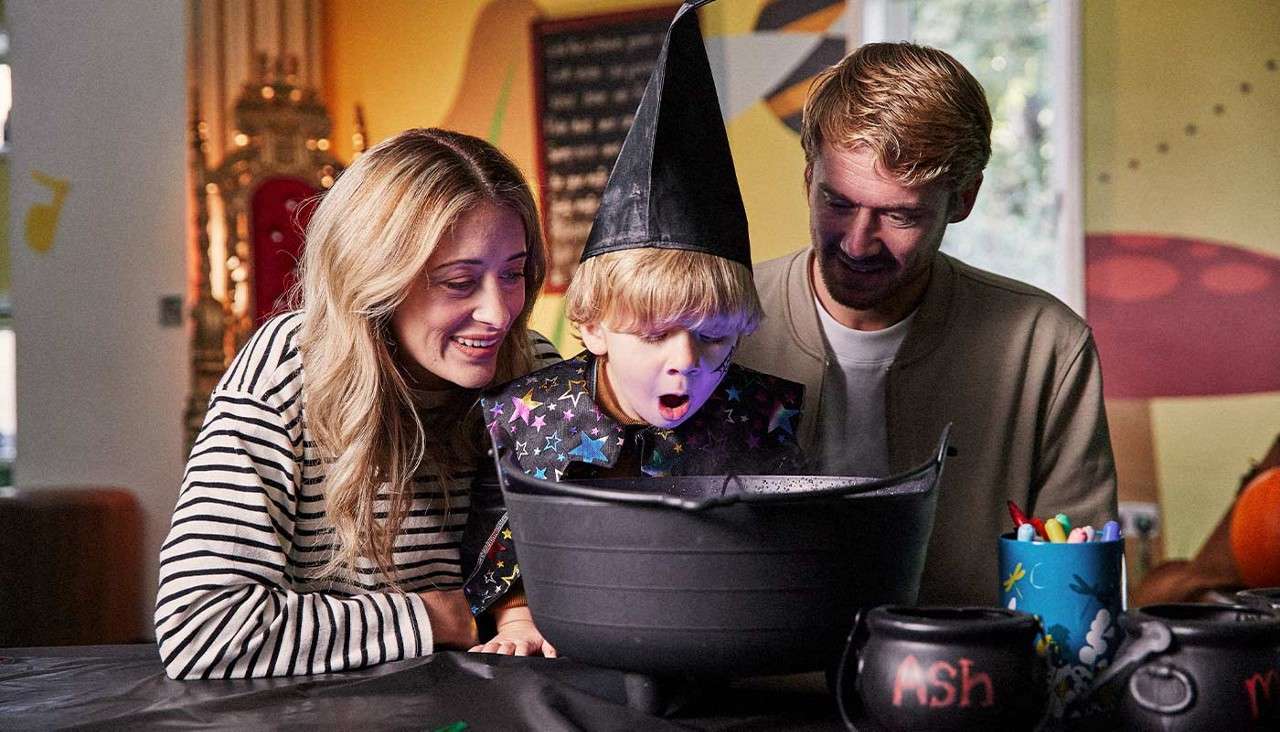 Child in a wizard hat peers into a black cauldron, gasping, while two adults smile beside him; Halloween-themed room with crafts and pumpkins. Visible text: "Ash" on a small cauldron.