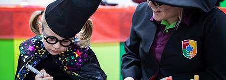 Child in a wizard costume writes with a marker, while an adult in a witch hat watches and smiles. They sit at a colorful table in a classroom-like setting.