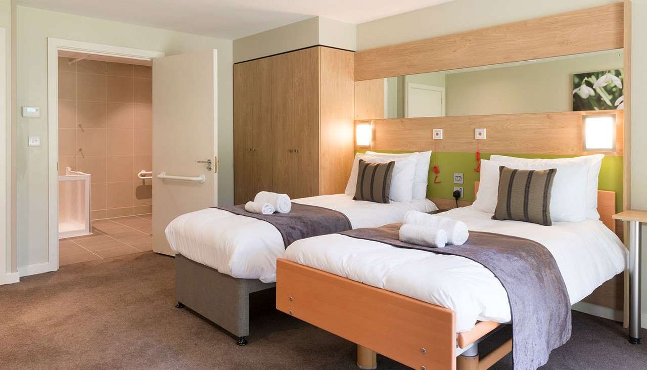 Twin beds sit neatly made with rolled towels, facing a wooden headboard with lights; a modern hotel room with wardrobe and an open door to an accessible tiled bathroom.