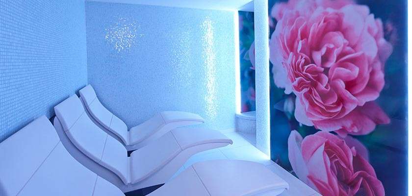 Curved white lounge chairs line a blue-lit tiled spa room, facing a wall with illuminated mosaic accents; a large mural of pink flowers covers one side, creating a tranquil setting.