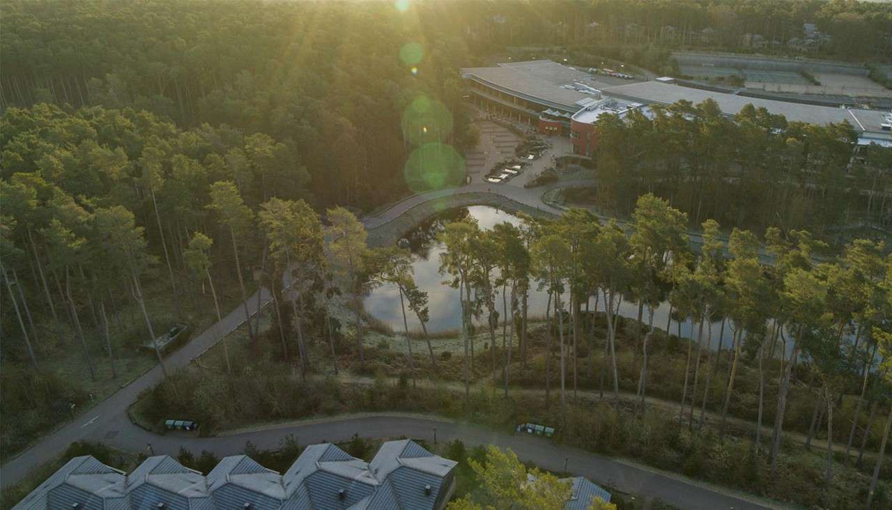 A resort complex borders a reflective pond, nestled within dense pine forest. Curved roads and paths encircle the water, parked cars line the entrance, and soft sun flares glow overhead.