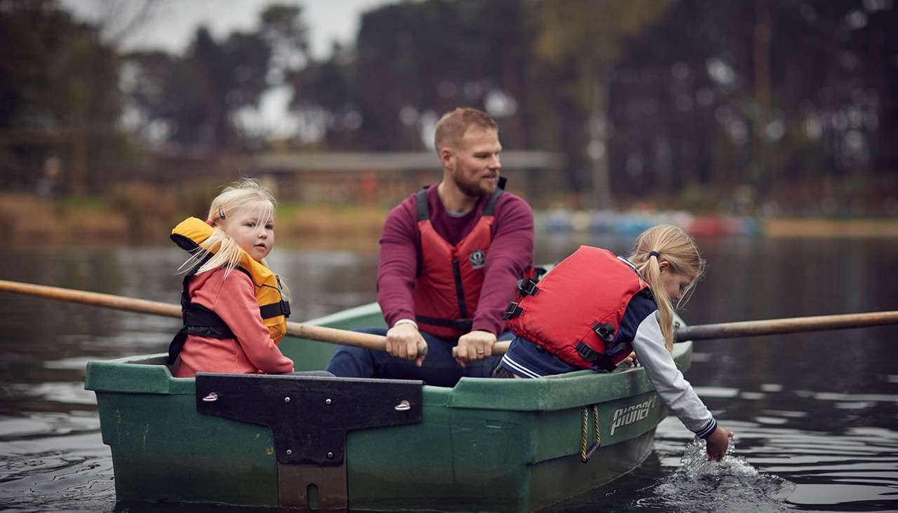 Green rowboat with life-jacketed man rowing and two children; one touches the water. On a calm lake bordered by trees. Text: Pioner.