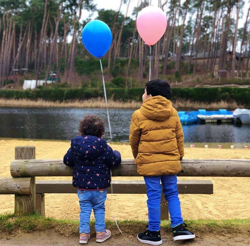 Two children stand against a wooden fence, holding blue and pink balloons, looking toward a small lake. Sandy shore, paddle boats, and tall pine trees create a calm outdoor setting.