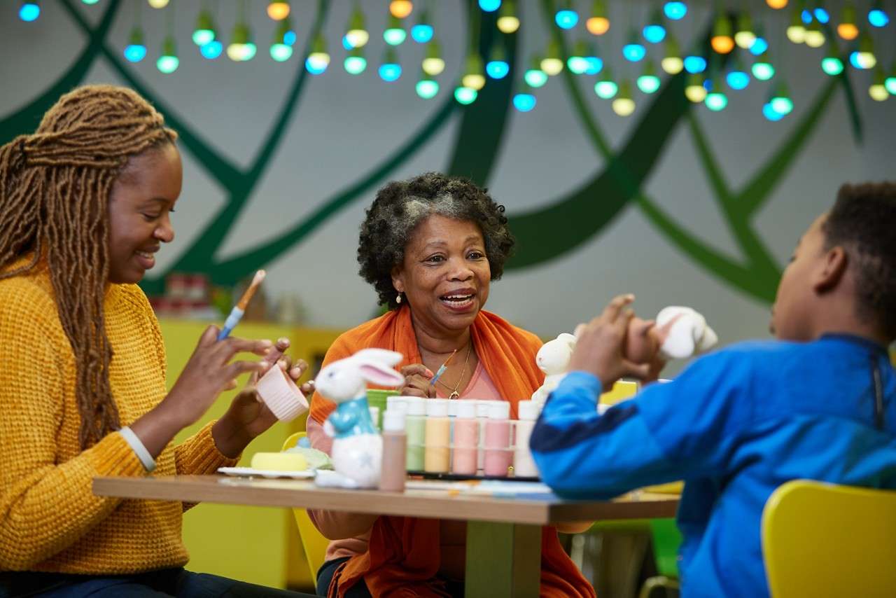 Three people paint ceramic bunny figurines and cups, chatting around a craft table, surrounded by pastel paints, brushes, yellow chairs, and colorful string lights against a green-patterned studio wall.