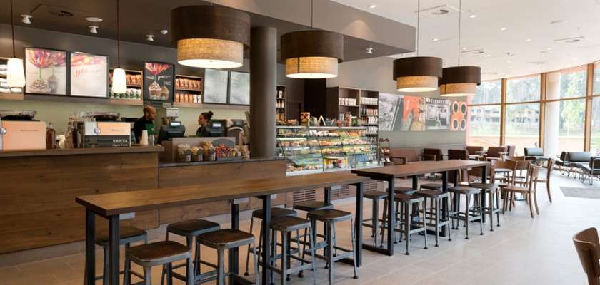 Coffee counter with two baristas prepares drinks, while a glass case displays pastries. Long communal tables and stools sit under pendant lights in a spacious, modern cafe with tall windows.