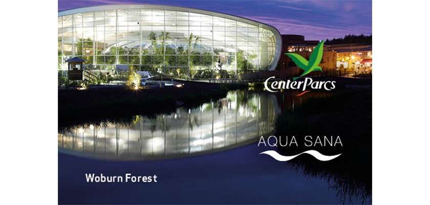 Glass leisure complex glows and reflects on a calm lake at dusk, surrounded by trees and nearby buildings. Text: CenterParcs, AQUA SANA, Woburn Forest.