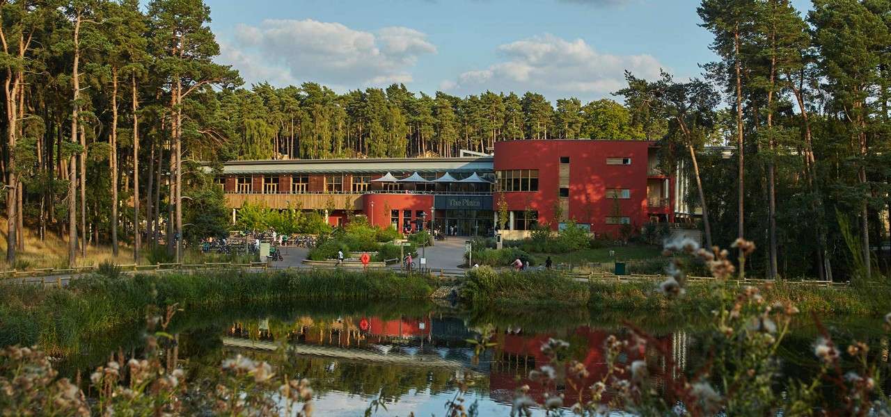 Red modern building faces a pond, reflecting in water while visitors walk and cycle nearby; sign above entrance reads "The Plaza". Tall pines surround the complex under partly cloudy sky.