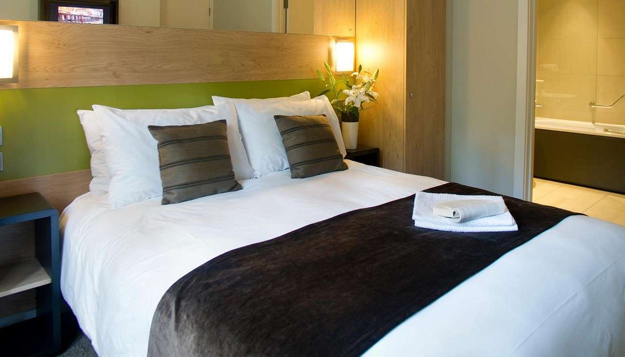 Bed holds folded towels and an open book on a dark runner; surrounded by white linens, cushions, bedside lamps, flowers, wood accents, and an adjoining bathroom with bathtub and handrail.