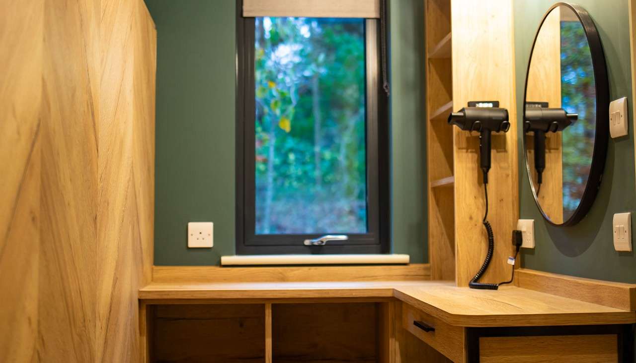 Wooden vanity anchors grooming; mounted hair dryers hang beside a round mirror; context: compact room with green walls, open shelves, power outlets, and a single window framing blurry trees outside.