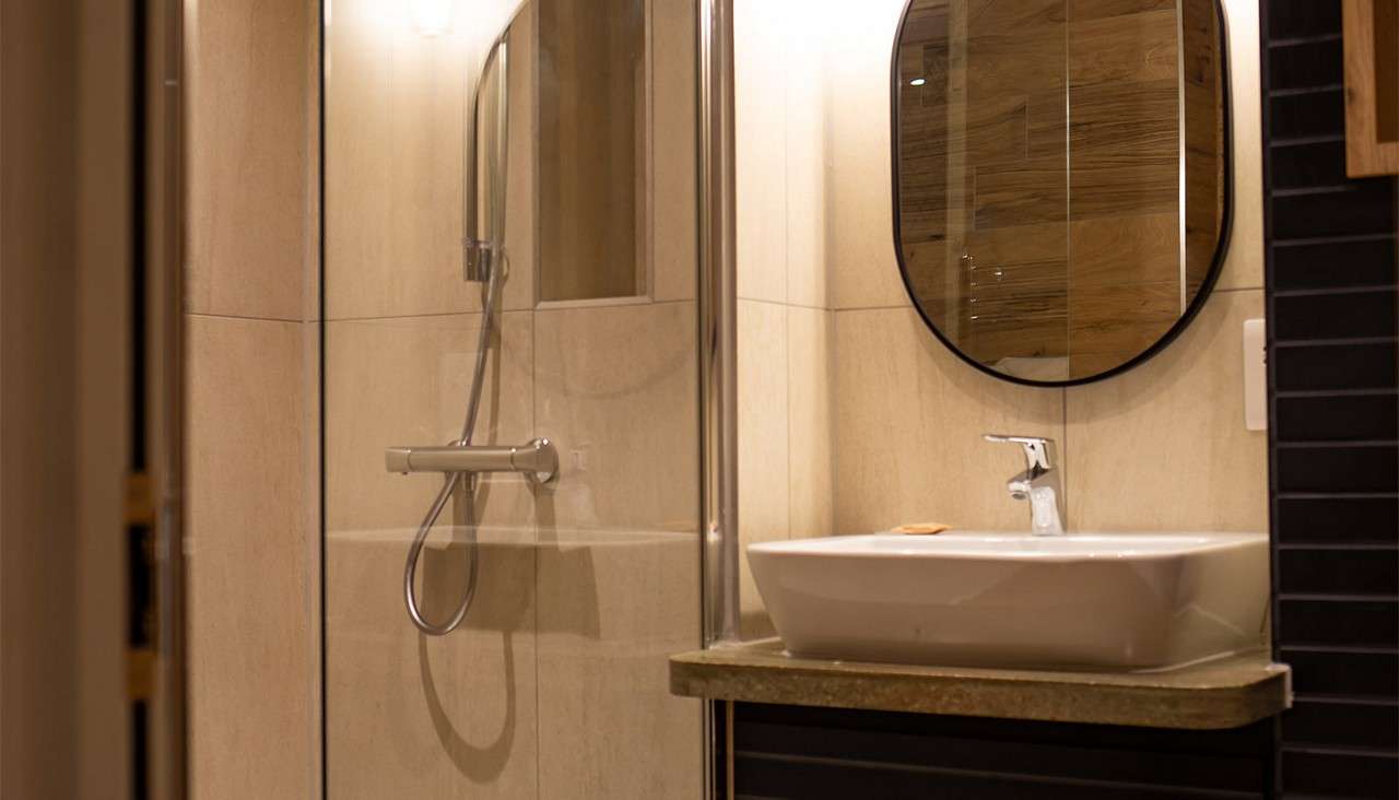 Bathroom sink rests under a round mirror, reflecting tiled walls, while a glass-enclosed shower with handheld sprayer and grab bar stands adjacent; warm lighting illuminates beige tiles and stone countertop.