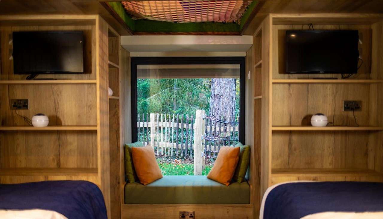 Window seat frames forest view, with green cushion and orange pillows. Surrounding context: wood-paneled cabin room with built-in shelves, two wall-mounted TVs, twin beds flanking, fence and trees outside.