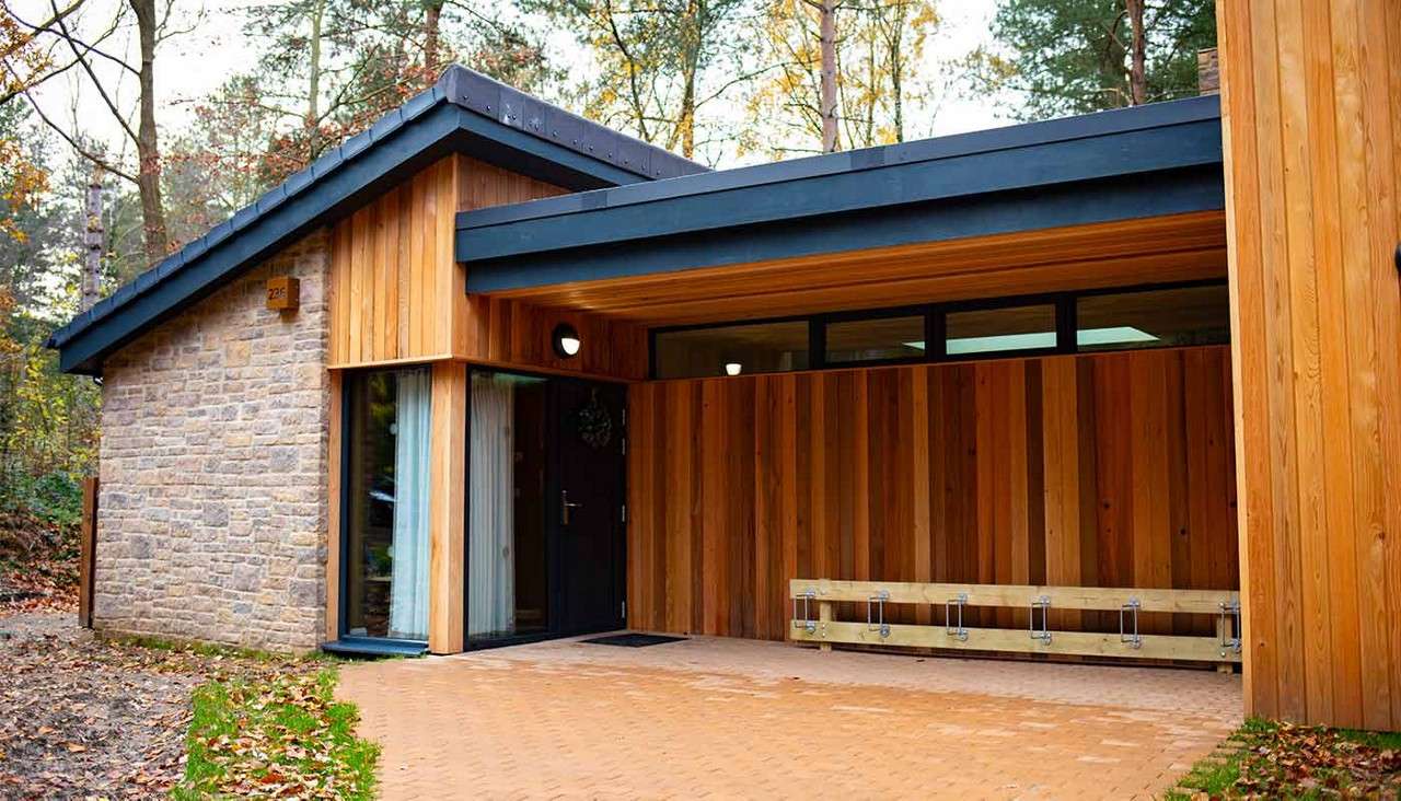 Modern timber-and-stone cabin sits under a deep overhang, sheltering a bench and dark entry door; narrow clerestory windows and sloped black-edged roofs face a paved drive in a forest.