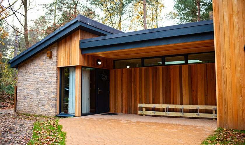 Wood-clad modern cabin shelters a covered entry with bench; stone accent wall and angled rooflines stand amid tall forest trees. Text: 21.