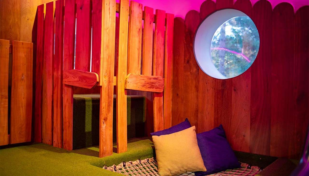 Cozy seating nook rests beneath pink lighting, featuring two pillows on a patterned mat. Wooden slat partitions frame carpeted steps, while a round window reveals trees, creating a playful, cabin-like interior.