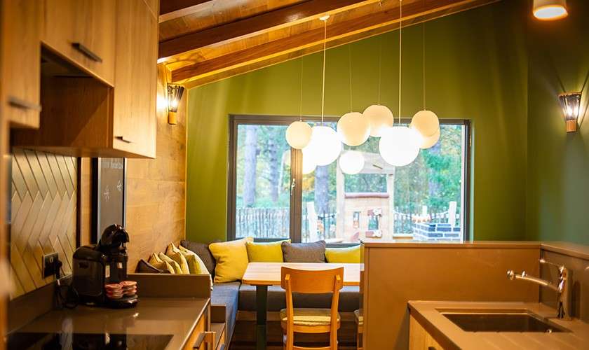 Pendant globe lights hang over a small dining table, illuminating a cozy kitchen-dining nook; wood cabinets, green walls, window overlooking trees, corner bench with cushions, sink and coffee machine nearby.