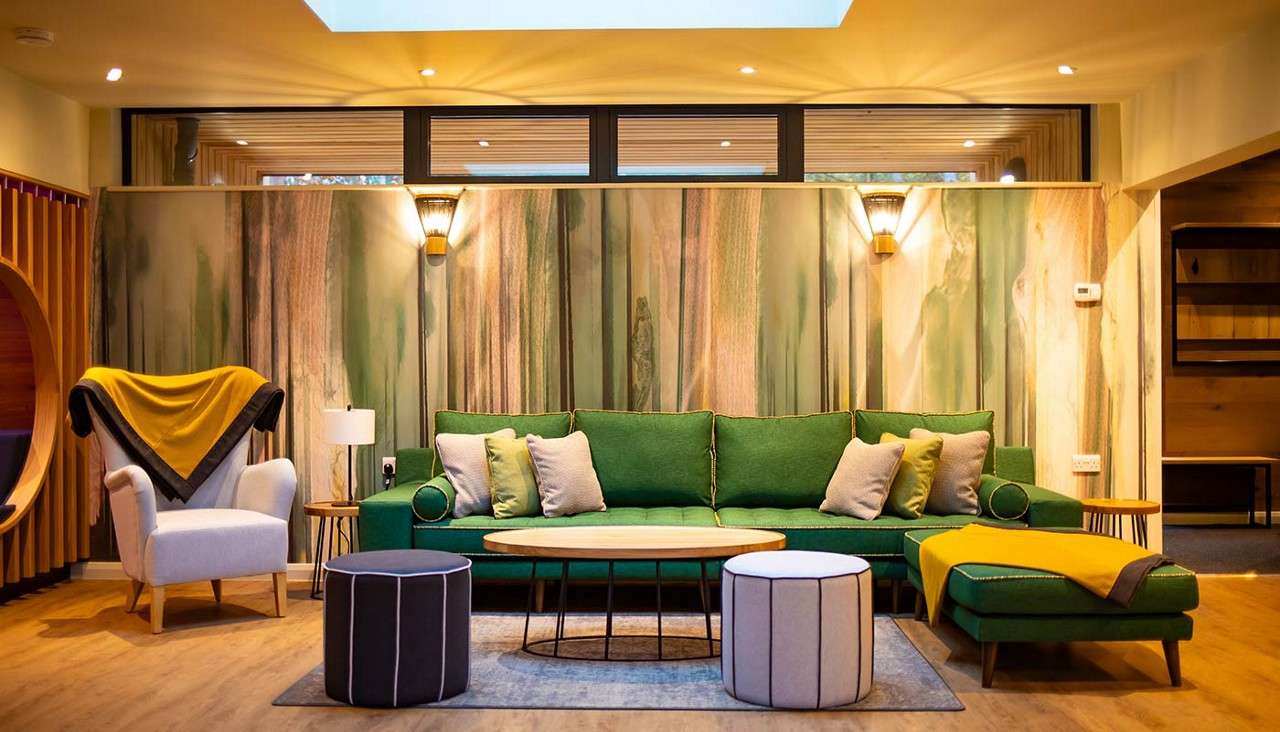 Green sectional sofa anchors the space, holding cushions and a yellow throw, facing round coffee tables and ottomans, in a warmly lit living room with wood floors, skylight, forest-pattern wall.