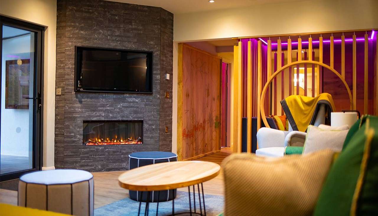 Electric fireplace glows beneath a wall-mounted TV, heating a contemporary lounge with round coffee table, ottomans, sofas, and blankets; a wooden slat divider with circular opening and purple lighting frames an adjacent hallway.