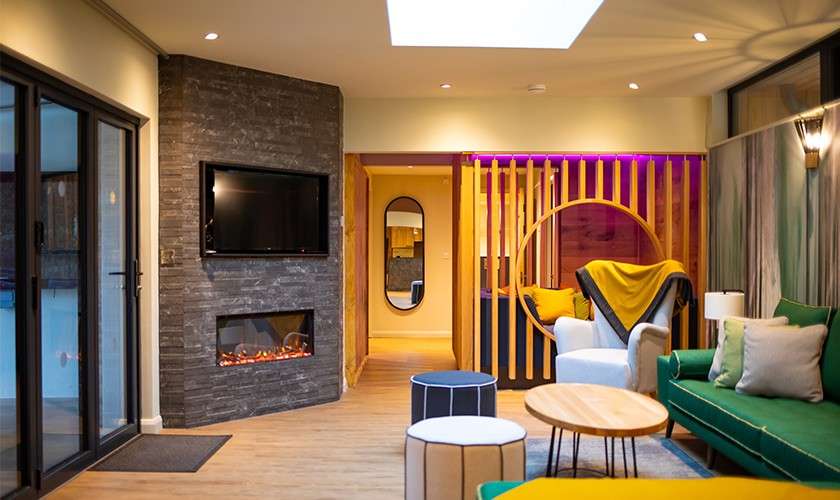 Contemporary living room hosts sofas and chairs arranged for conversation, facing a TV above an electric fireplace; skylight, sliding doors, small tables, and a wooden partition with purple backlighting surround.