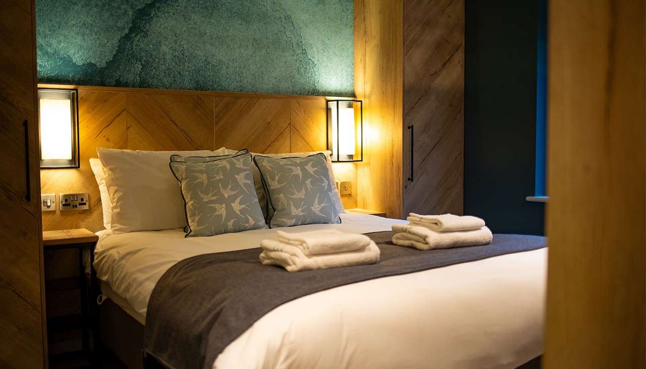 Bed holds folded towels atop a gray throw under warm wall lights. Surroundings include a chevron wood headboard, teal artwork, bird-print cushions, bedside tables, and wardrobe against a dark accent wall.