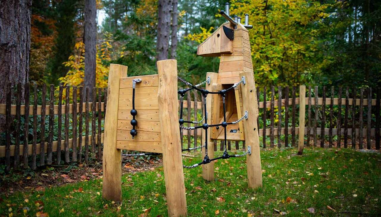 Wooden animal-shaped playground structures connect via rope-and-chain climbing tunnel for children; set on grass within a rustic fenced area surrounded by trees with autumn foliage.