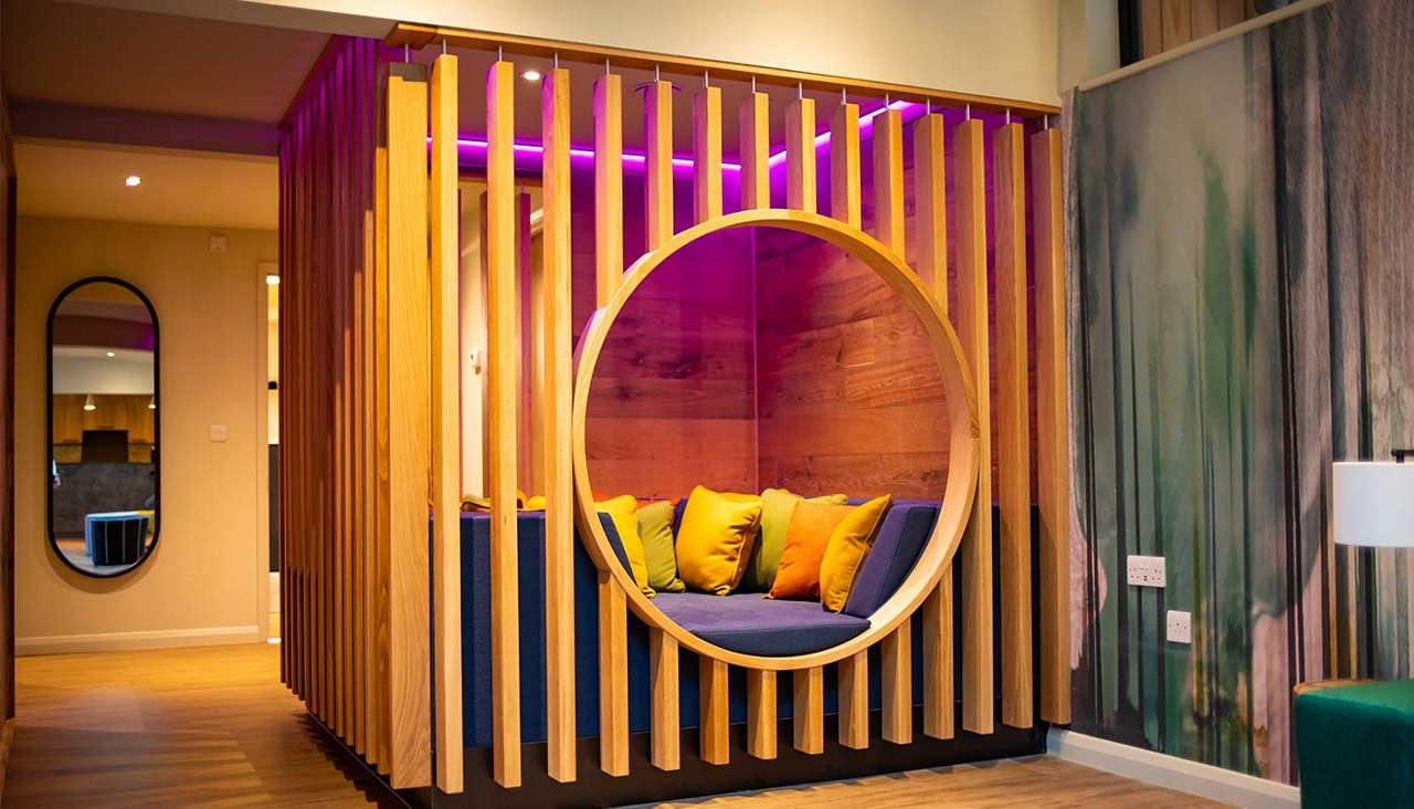 Circular wood-framed seating nook holds blue bench and colorful cushions, illuminated by purple strip lights, within a slatted timber booth; contemporary lounge surrounds with mirrors, wood flooring, and wall art.