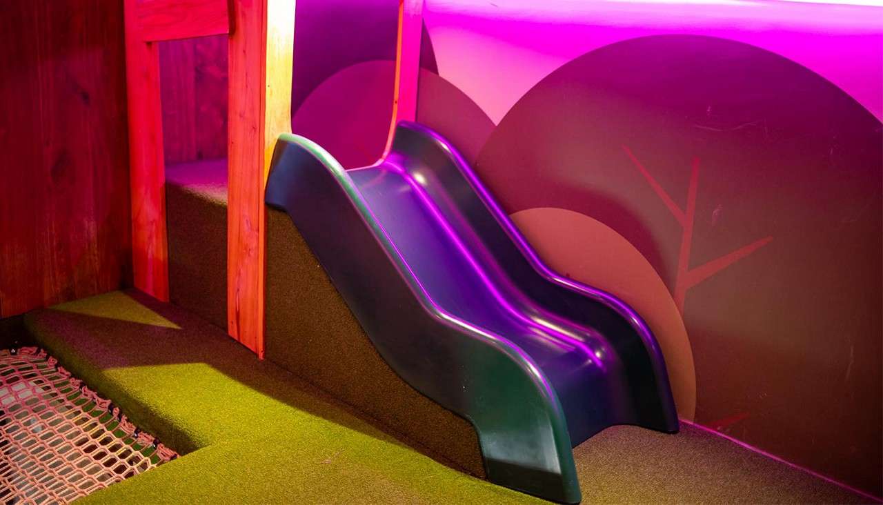Plastic children's slide descends between wooden supports, glowing under purple light. Surrounding context: indoor play area with green carpeted steps, safety netting, and a wall mural of stylized trees.