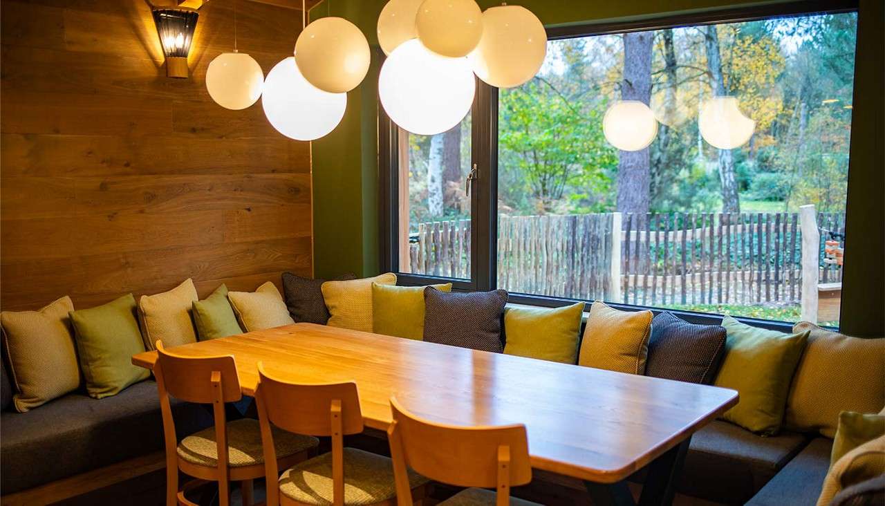 Pendant globes illuminate a wooden dining table in a cozy nook with bench cushions; large window overlooks trees and a rustic fence.
