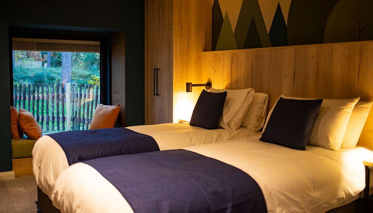 Twin beds rest side-by-side, topped with white linens and navy throws, under warm lamplight. In a cozy wood-paneled bedroom, a window seat faces a fenced garden and geometric wall mural.