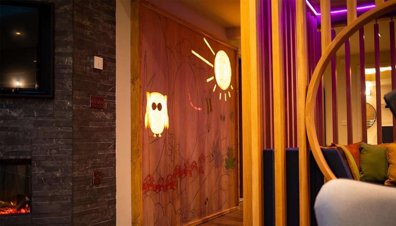 Glowing owl and sun illuminate a wooden mural. In a modern lounge, wood slats and a circular chair frame colorful cushions, beside a stone fireplace and purple LED accents.