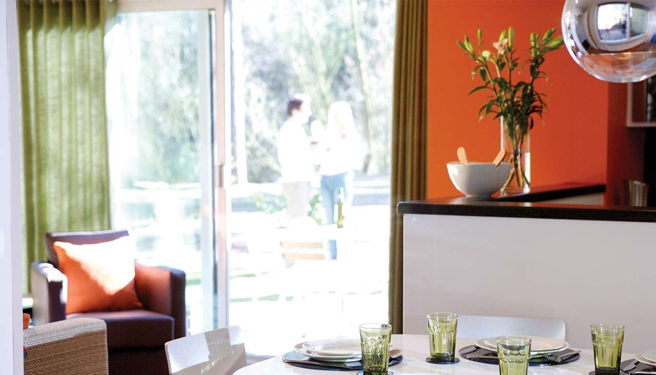 Dining table holds plates and green glasses, set for four; sunlight streams in as two people converse outside through an open sliding door; nearby counter displays vase and bowl.