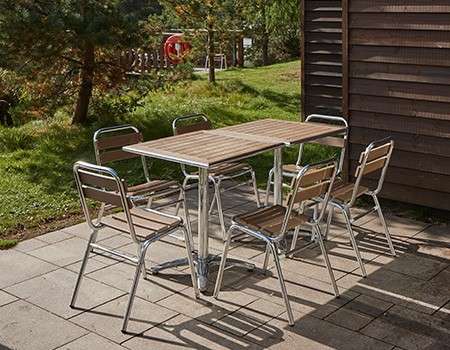 Outdoor dining set—rectangular wooden table with six metal-framed chairs—stands unused, casting shadows on a paved patio beside a wooden-sided building, with grassy lawn, trees, and sunlight beyond.