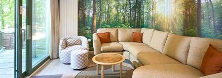 Curved beige sectional anchors the room, facing a round wood coffee table. Nearby, patterned chair and ottoman sit on a rug, beside sliding glass doors; forest mural brightens the wall.