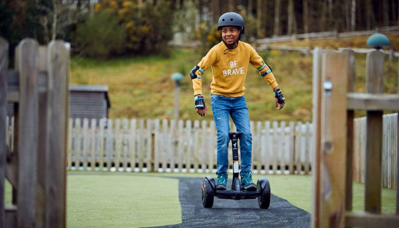 Child wearing helmet rides a two-wheeled self-balancing scooter along a narrow paved path in a playground; wooden fence and trees behind. Sweatshirt text: BE BRAVE.