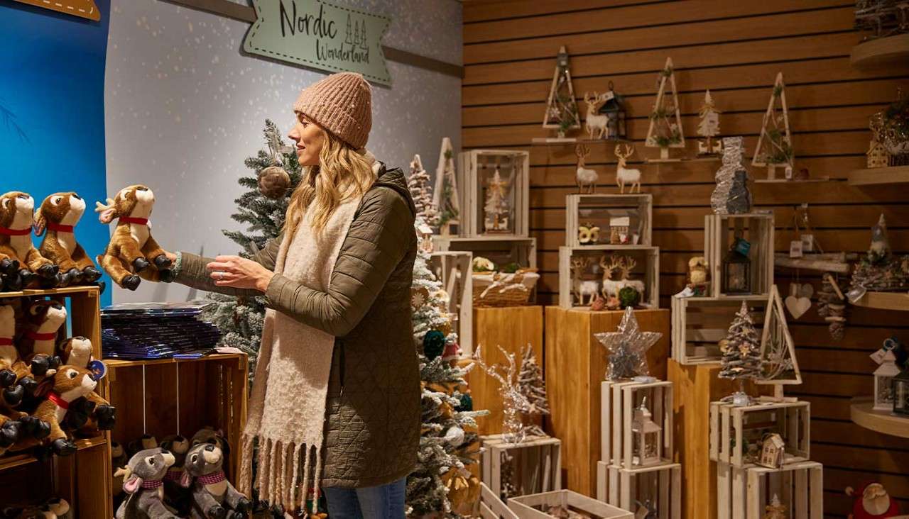 Shopper holds a plush reindeer, examining it. Context: cozy Christmas shop with wooden crates, small trees, ornaments, and a sign reading “Nordic Wonderland”.