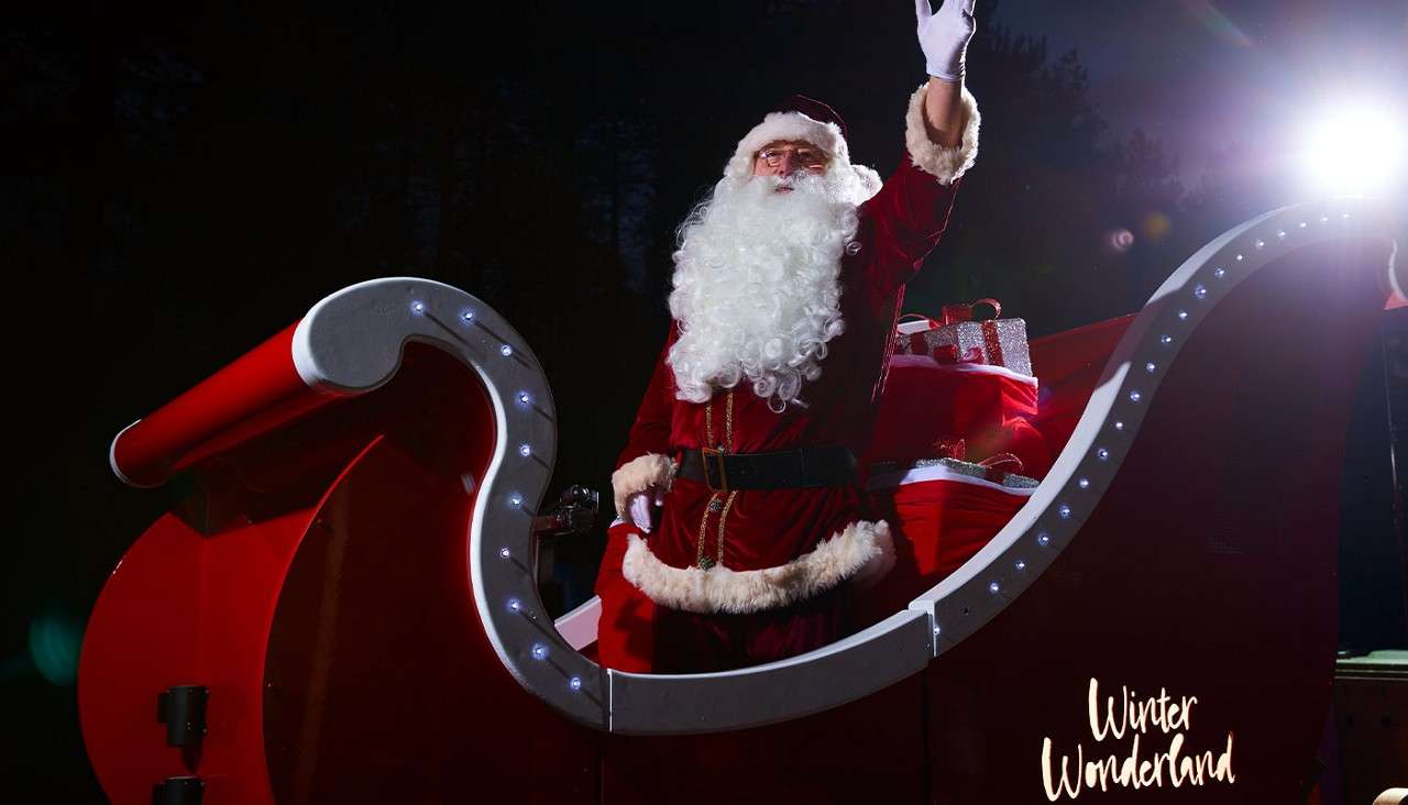 Person dressed as Santa waves from a red, light-trimmed sleigh, with wrapped gifts behind, under a night sky and bright spotlight. Text on lower right reads: Winter Wonderland.