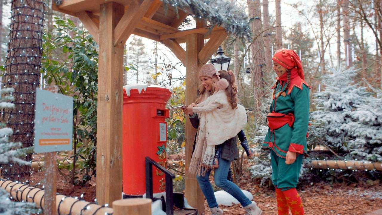 Mother and child post a letter into a large red mailbox; an elf-costumed attendant watches; snowy decorated woodland with wooden shelter and lights. Visible sign text: Don't forget to post your letter to Santa. Smaller text illegible.