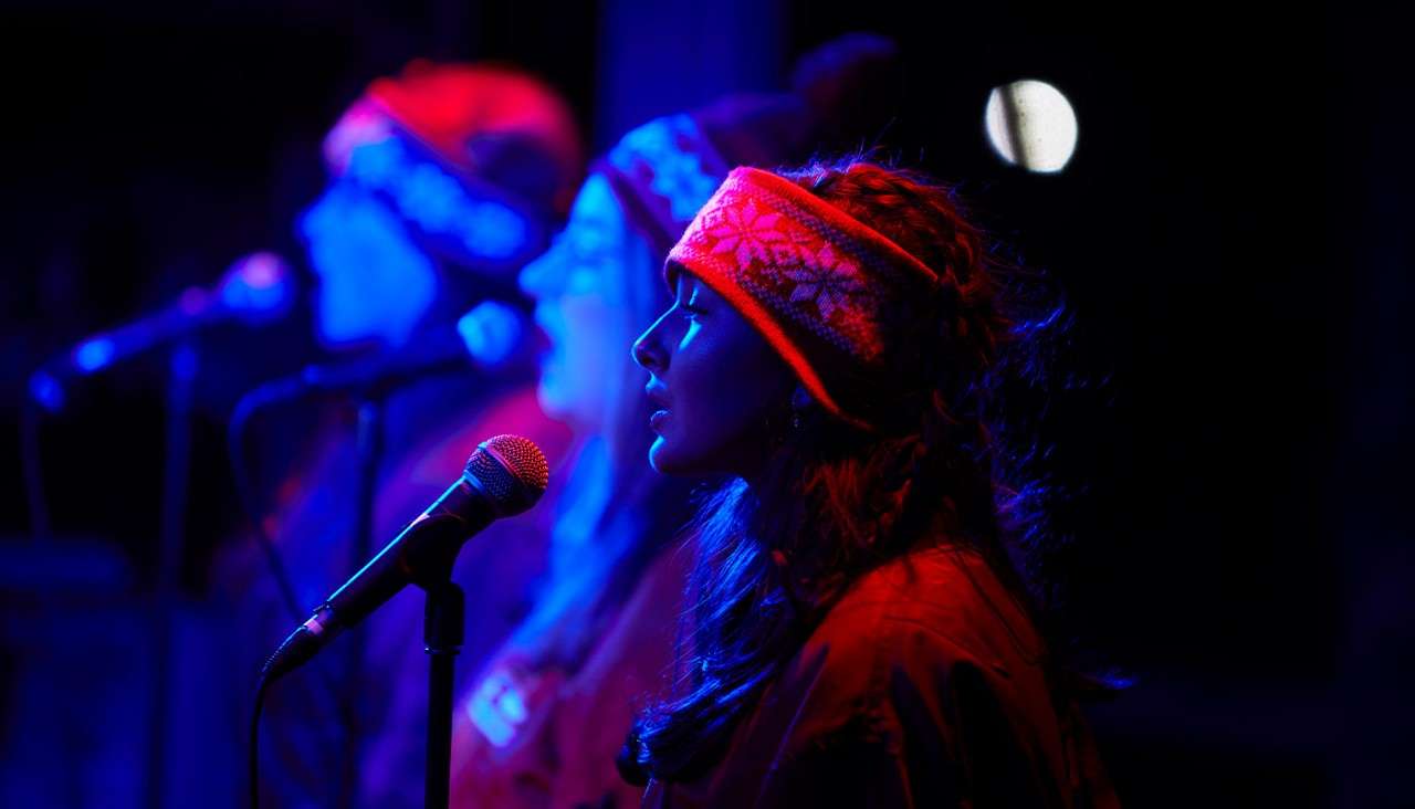 Three bundled singers perform into microphones, profiles lit by red and blue stage lights, wearing patterned winter headbands, standing closely on a dark stage with blurred background and spotlights.