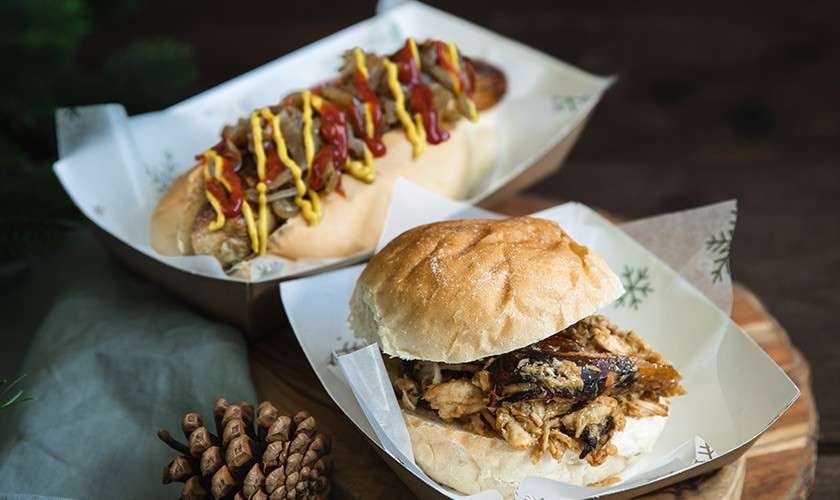 Two sandwiches sit in paper trays: a hot dog topped with mustard, ketchup, and onions, and a pulled meat bun. They rest on a wooden surface beside pinecones and greenery.
