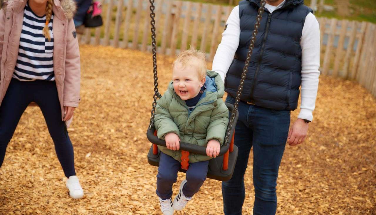 Child in a green jacket smiles while riding a bucket swing, gently pushed by two adults, in an outdoor playground with wood-chip ground and a wooden fence.
