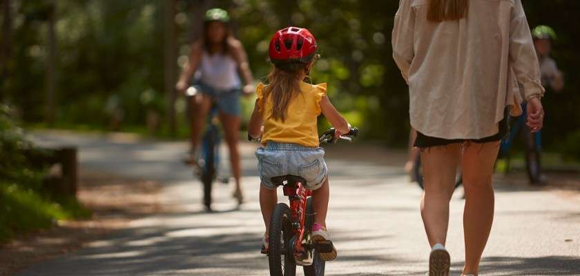Child on small red bicycle rides forward, wearing a red helmet; adult walks beside. In the background, other cyclists approach along a sunlit, tree-lined park path.