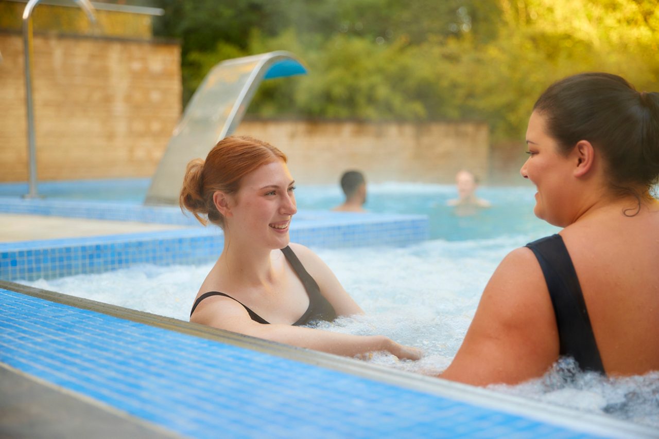 Two adults relax, chatting in a bubbling outdoor spa pool; steam rises, tiled edges and a curved waterfall feature behind, with others bathing in the background.