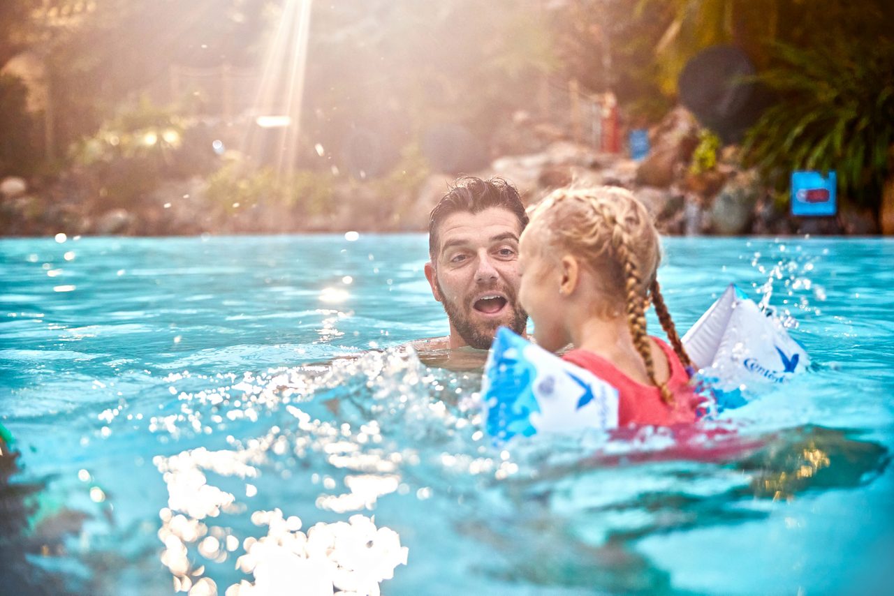 Two people swim and interact in a sunlit pool; a child with inflatable armbands faces an adult; surrounding tropical foliage and rocks border the clear water.