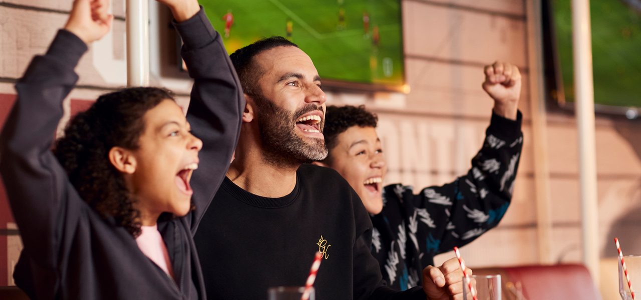 Three people cheer with raised arms, smiling and shouting, while seated at a table with striped-straw drinks; televisions behind them display a soccer match in a casual sports bar setting.