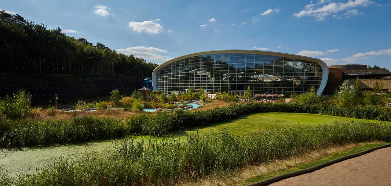 Curved glass leisure building reflects sunlight, enclosing water slides, while landscaped gardens, a lazy river, reeds, and a pathway spread in front, bordered by a wooded hillside under blue sky.