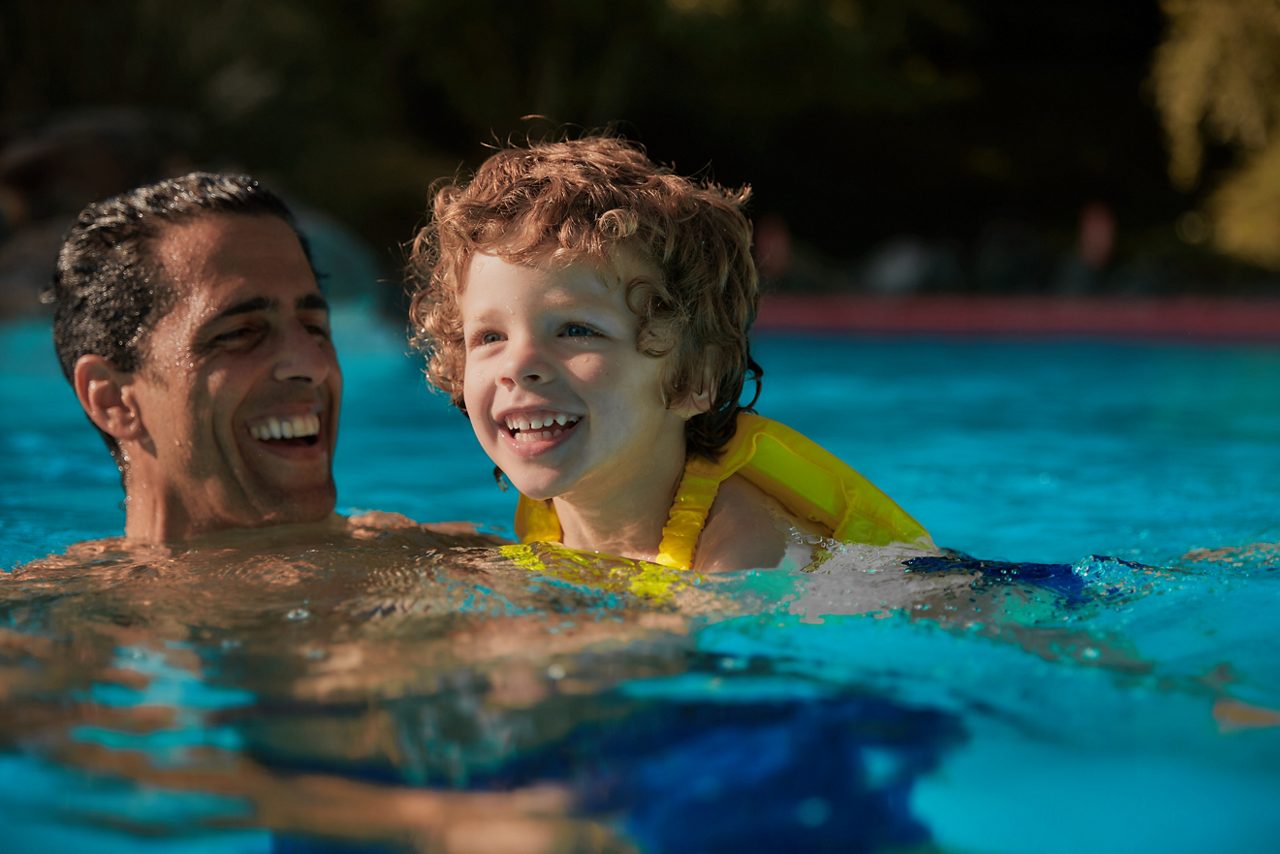 Young boy wearing a yellow buoyancy aid, as he swims with an adult.