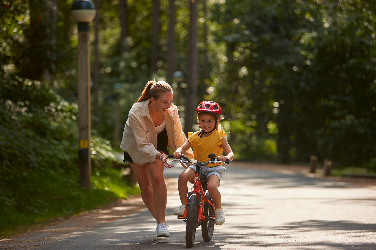 A mother helping her daughter ride a cycle through the forest.