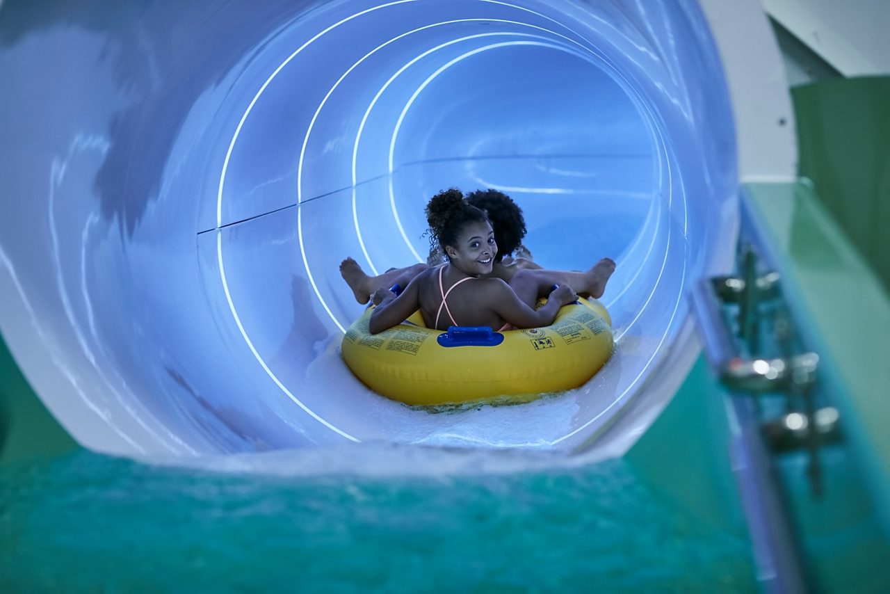 Two young people sitting in an inflatable raft going down a tube slide.